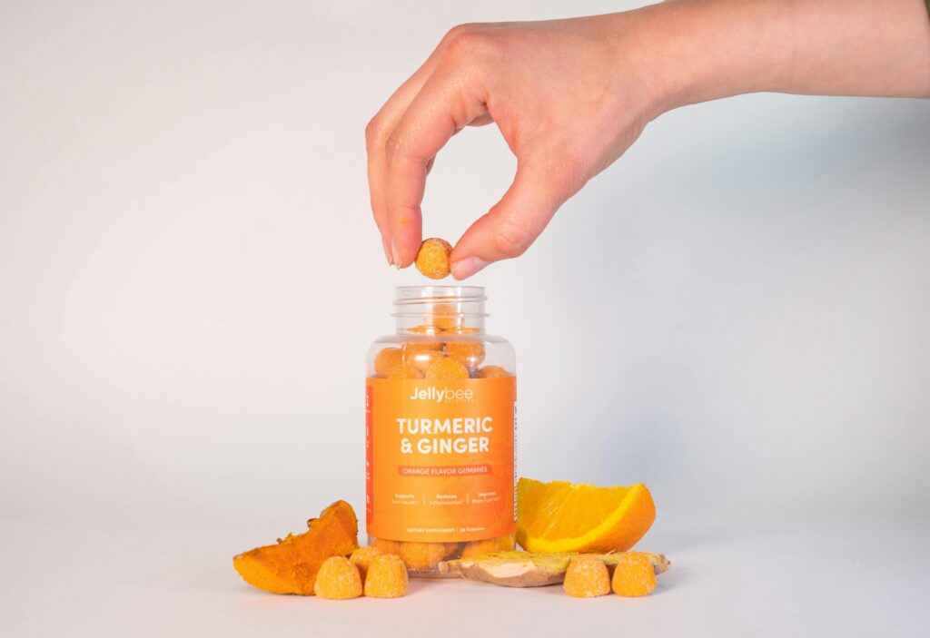 pexels-photo-8940751-8940751 Close-up of a hand taking a turmeric and ginger gummy from a bottle, surrounded by ingredients.