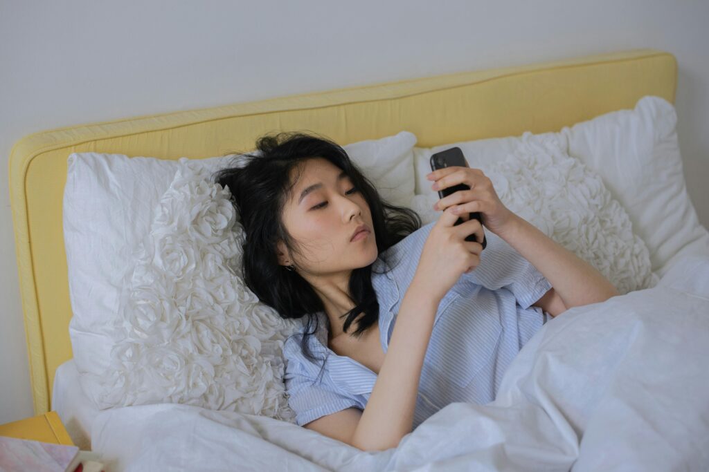 pexels-photo-7341879-7341879 Asian woman comfortably lying in bed, using her phone in a serene bedroom setting.