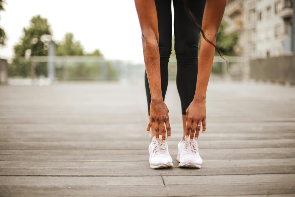 pexels-photo-3769002-3769002 From below crop slender female athlete in sportswear and white sneakers doing standing forward bend exercise for stretching body on wooden floor of street sports ground against blurred urban environment in daytime