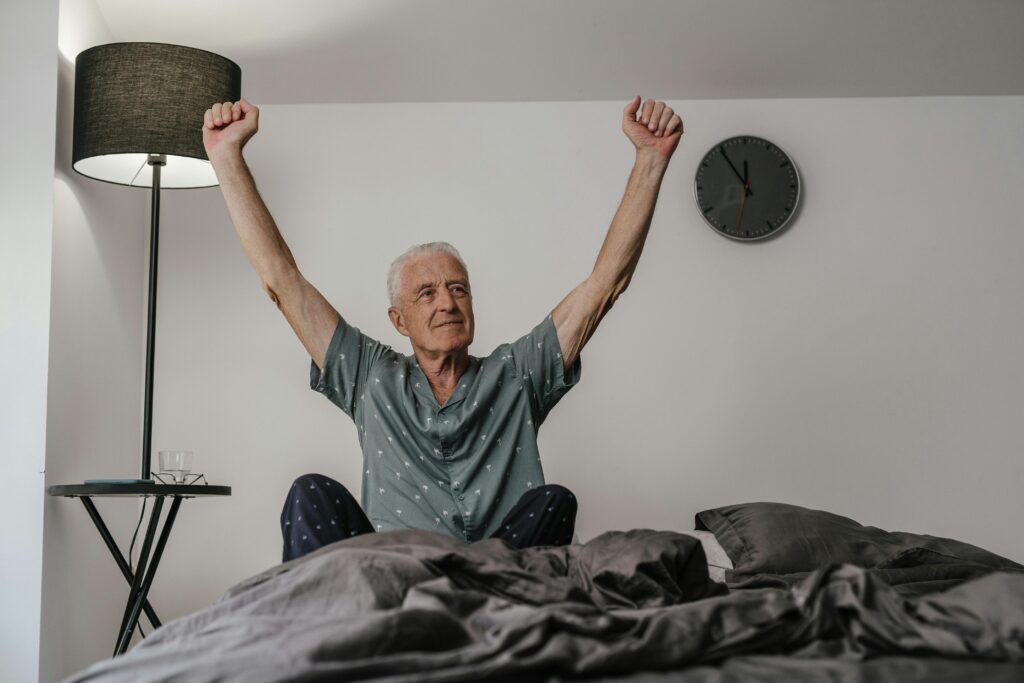 pexels-photo-8899494-8899494 Senior man in pajamas waking up with arms stretched in a cozy bedroom setting.